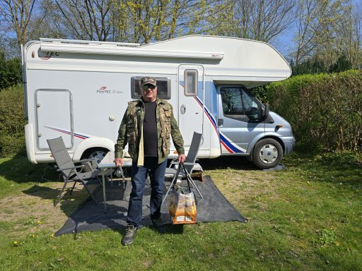 Mann mit rotem Koffer vor einem Wohnmobil, mehrere Stühle und eine Matte im Freien.
